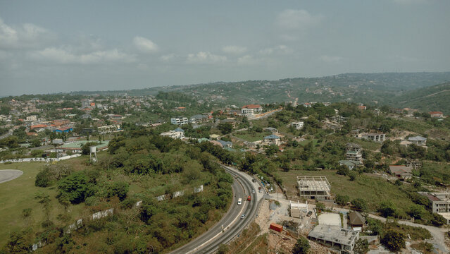 Aerial view of winding roads cutting through lush green hills and scattered buildings under a vast sky, Aburi, Eastern Region, Ghana.