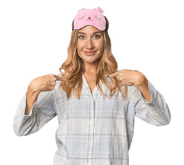 Blonde in pajamas with sleep mask surprised pointing with finger, smiling broadly.