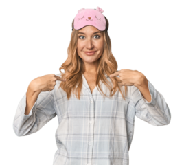 Blonde in pajamas with sleep mask surprised pointing with finger, smiling broadly.
