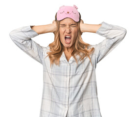 Blonde in pajamas with sleep mask covering ears with hands trying not to hear too loud sound.