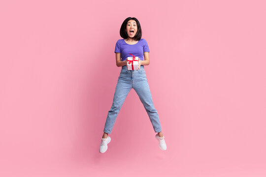 Wow. Full length pportrait of jumping high amazed surpised asian young woman holding wrapped gift box with present, celebrating holiday isolated over pastel pink studio background