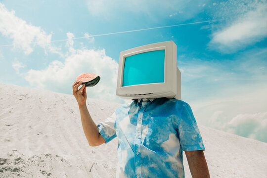 Person with CRT monitor head holding fruit on sandy dune in summer