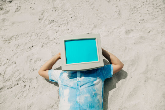 Person lying on sand with vintage computer monitor on head as creative metaphor