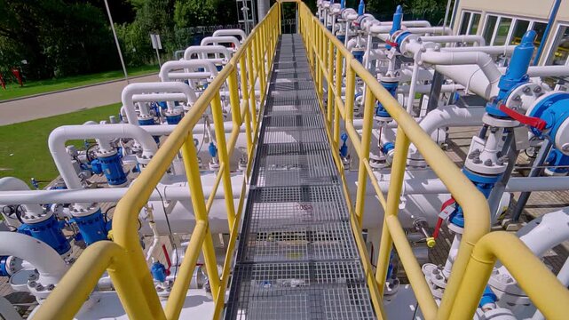 Metal catwalk between rows of pipes and blue valves at a modern natural gas pumping station and transportation facility in Latvia, revealing the infrastructure - A dolly-back reveal shot