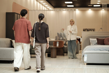 Young adult man and woman walking together toward woman standing and gesturing in furniture store...