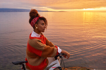 Outdoor portrait of happy african woman admiring sunset over the lake during bike ride