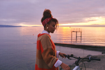 Outdoor portrait of happy african woman admiring sunset over the lake during bike ride