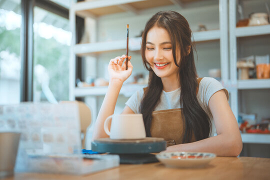 Happy asian woman thinking of creative cup painting and product design ideas with handmade ceramic mug at pottery studio – smiling small business owner in artisan clay workshop interior