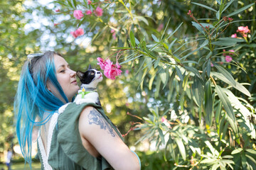 Woman with blue hair holding cat smelling pink flowers in a garden