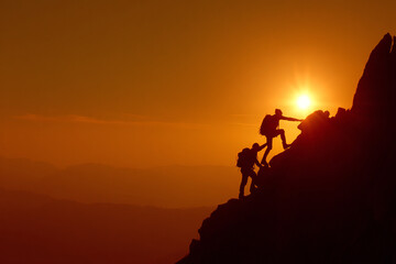 Two climbers ascend a steep rock face, with one helping the other, silhouetted against an orange sunset.