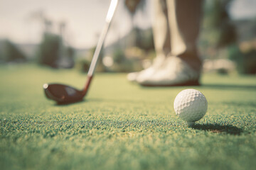 A close-up of a golf ball on a tee with a golf club and a player's feet in the background.