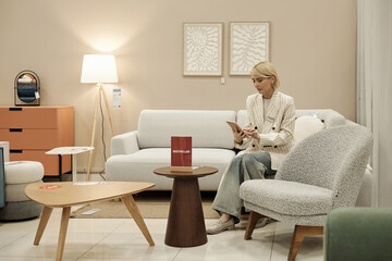 Caucasian woman sitting on chair reading brochure in furniture store showroom surrounded by modern...