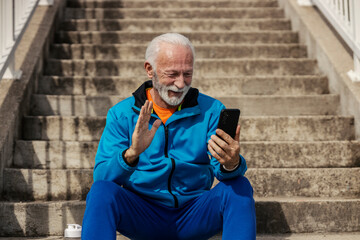 Portrait of senior sportsman waving at cellphone during video call while sitting on staircase outdoors.