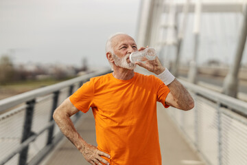 Tired thirsty senior sportsman in shape is drinking water on a bridge.