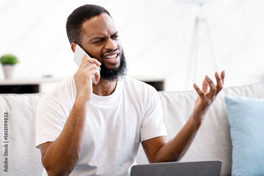 Poster discontented black guy talking on cellphone sitting at laptop having computer problem at home. displ