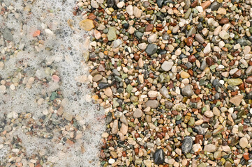 Colorful sea pebbles scattered along shoreline, with wave gently washing over them. multicolored beach pebbles and fine gravel at edge of sea. Gentle wave foam touches stones. marine background.