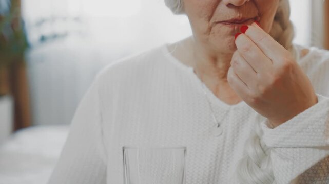 Happy elderly senior woman sitting on bed eating taking vitamins pill holding glass water drinking. Lady pension age cropped face closeup mouth lips, old age health care. Fish oil. hormonal therapy