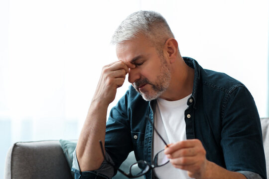 Tired senior man removing glasses, rubbing nose bridge, having tired eyes syndrom after working, reading or using gadgets. Matured man sitting on couch in living room at home, copy space - Powered by Adobe