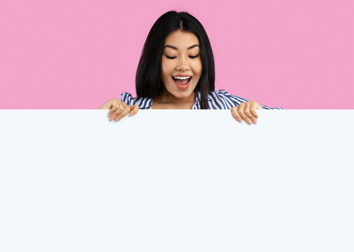 Fototapeta Wow. Excited asian female standing behind white empty board for advertisement over pink background. Cheerful lady with open mouth looking down at big blank advertisement placard, empty space for text