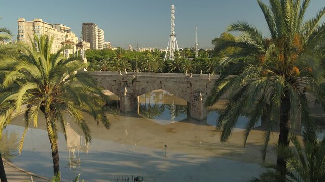 Ascending aerial drone shot from Turia Gardens in Valencia featuring the Puente del Mar, palm reflections in the lake, and a Ferris wheel in the distance