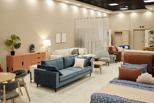 Modern furniture showroom displaying various sofas, beds, chairs, tables and decor items arranged in spacious interior with bright lighting, no people visible, clean tiled floor