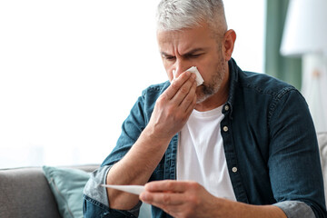 Sick elderly man sneezing and checking body temperature, sitting on sofa in living room, copy space. Senior man holding thermometer, suffering from fever, got cold, flu or coronavirus