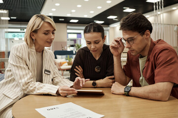 Caucasian young adult woman showing digital tablet to young adult woman and young adult man sitting...