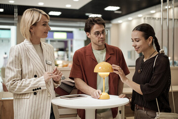 Caucasian woman assisting young adult Caucasian man and young adult woman examining modern table lamp together in furniture store showroom