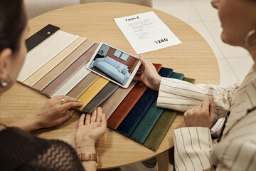 Two Caucasian women sitting at round table discussing fabric samples while holding tablet...