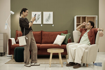 Young adult Caucasian woman standing and photographing young adult Caucasian man sitting on...