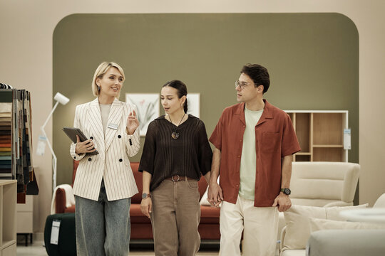 Woman holding tablet guiding young adult woman and young adult man through furniture store, group walking together and interacting