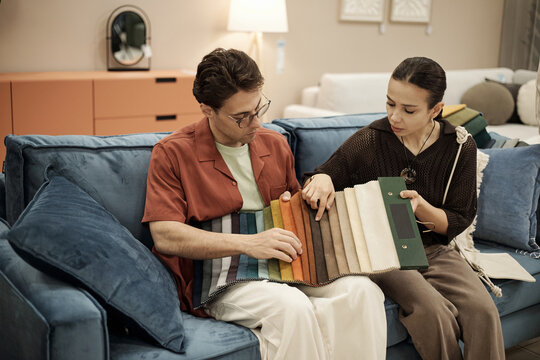 Caucasian young adult man and Caucasian young adult woman sitting on sofa examining fabric swatches together, discussing material options in furniture store setting