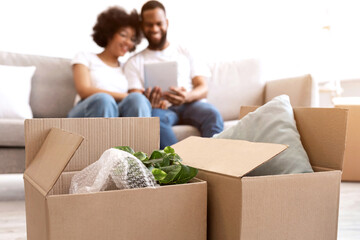 Moving Boxes Standing On Floor While Happy Black Couple Using Digital Tablet Packing For A House Move Sitting On Sofa Indoor. Family Housing, Real Estate, New Apartment Concept. Selective Focus