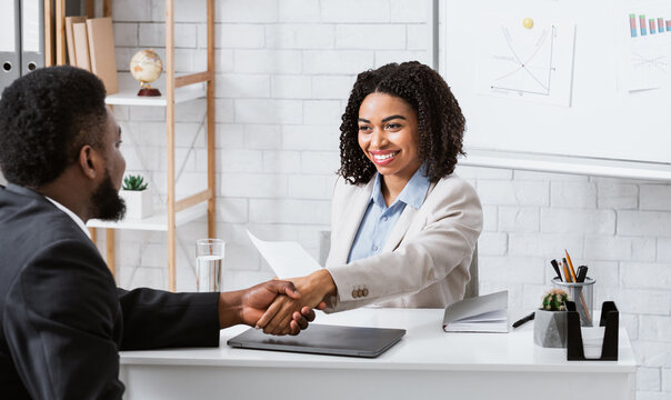 Positive female personnel manager and black job applicant shaking hands before employment interview at company office. Specialist headhunter communicating with vacancy candidate