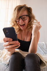 Excited woman celebrating success on smartphone in bed