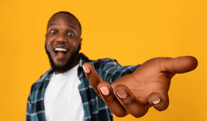 Happy african american guy outstretching hand, holding something invisible on his huge palm, giving it away or asking for something, orange studio background, collage, panorama with copy space
