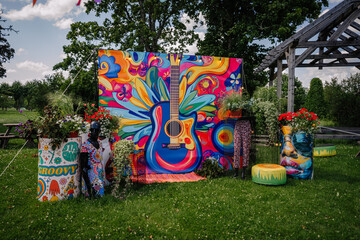 Vibrant hippie-themed outdoor decoration featuring a large colorful guitar backdrop, mannequin, plants, and psychedelic artwork on barrels and cushions.