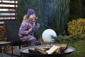 An eight-year-old girl roasts marshmallows on an autumn day near a country house.  Family leisure with kids at autumn.