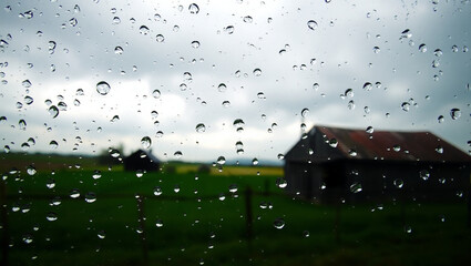 Raindrops running down a window pane with blurred countryside outside with Green fields, a rustic barn, and a cloudy sky