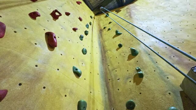 Low-angle upward view of an indoor climbing wall featuring red and green climbing holds and a dual rope top-rope system. Ideal for sports promos, climbing tutorials, advanture content, or fitness.