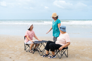 Senior women enjoying and talking while sitting on folding chairs on tropical beach. Asian female...