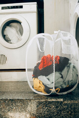 White laundry basket filled with colorful clothes placed in front of a washing machine. Household chore scene with laundry equipment. Household chores, laundry, daily life concept.