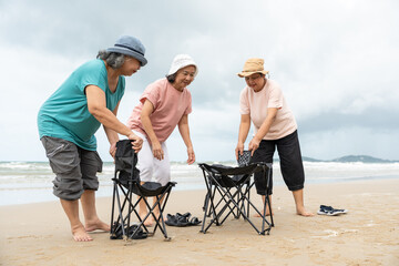 Senior women preparing folding chairs to relax and enjoy their time by the sea on beach. Asian...