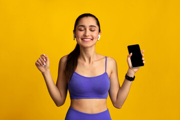 Enjoying Favorite Music For Training. Portrait Of Smiling Happy Young Sportswoman Wearing Wireless Earbuds Listening To Workout Playlist Holding Mobile Phone With Blank Empty Screen, Mockup Template