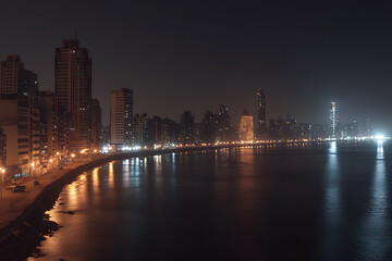 Fototapeta premium City skyline at night with waterfront. Illuminated buildings reflect in the calm water. A serene view of urban life meeting the sea.