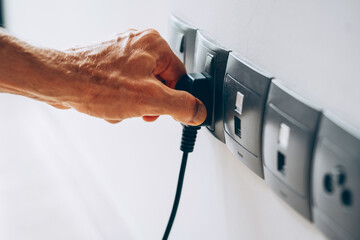 Hand plugging a black electrical plug into a wall socket. Several sockets are aligned on a white wall in a modern interior.