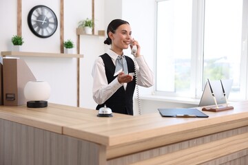 Smiling receptionist talking with client by phone at reception desk in hotel