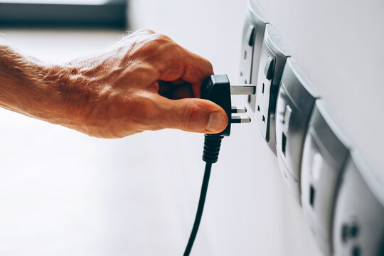 Hand plugging a black electrical plug into a wall socket. Several sockets are aligned on a white wall in a modern interior.