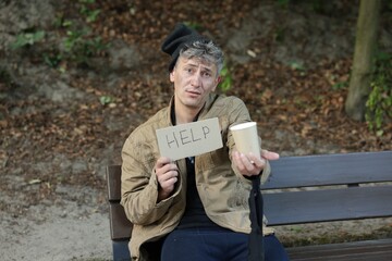 Homeless man with sign Help and paper cup begging for money on bench in park