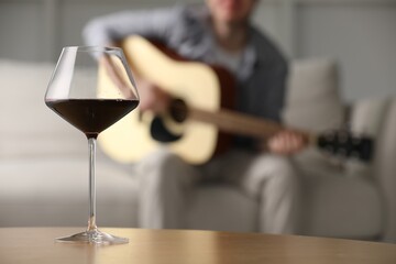 Glass with red wine on table and man playing guitar indoors, selective focus
